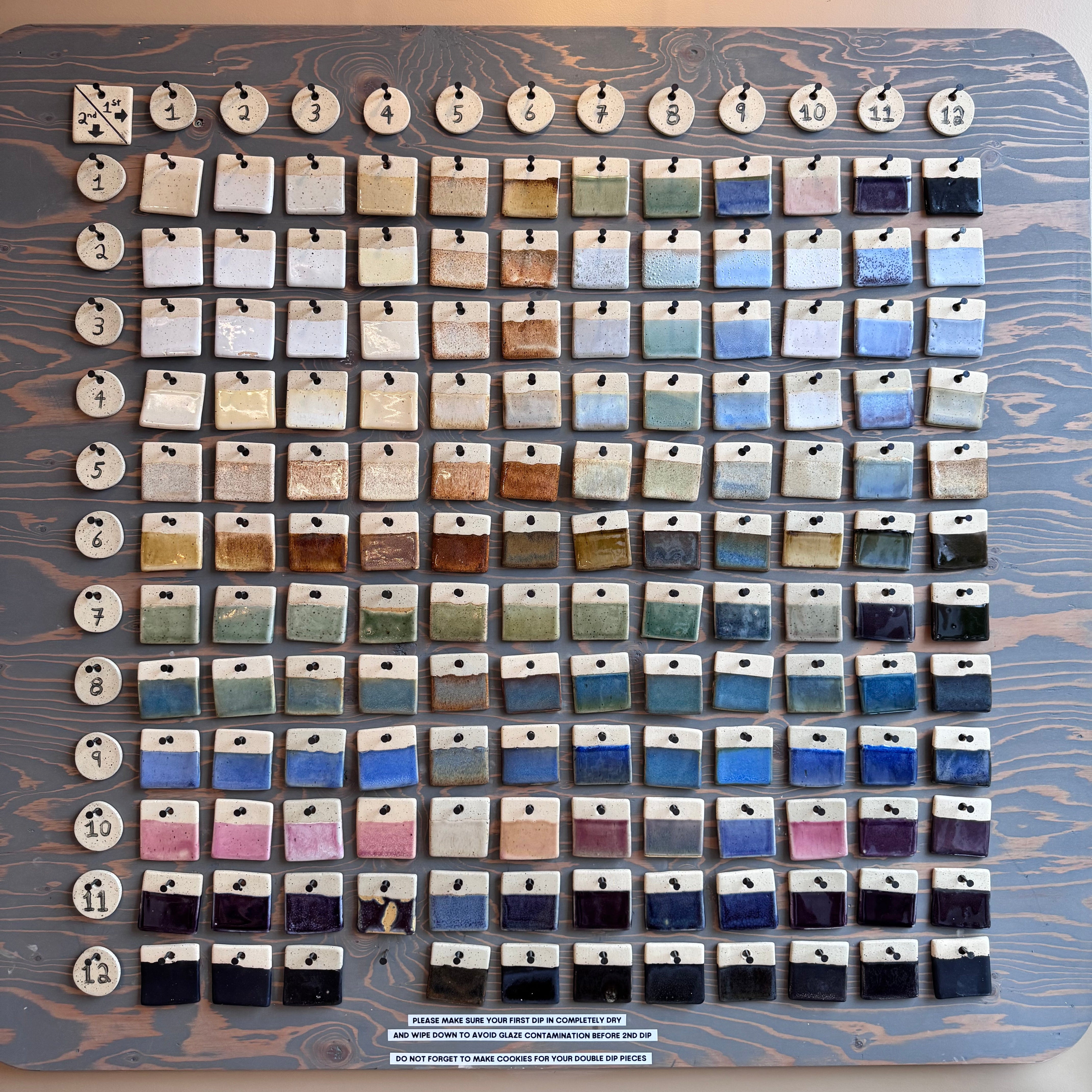 A large display board showing many small rectangular glaze test tile samples arranged in rows, labeled with numbers around the top edge. The board has a woodgrain finish and is photographed indoors against a beige wall.