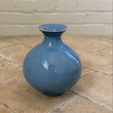 A glossy blue ceramic vase with a rounded bulbous body and a flared neck, standing on a stone-tiled floor against a white brick wall.