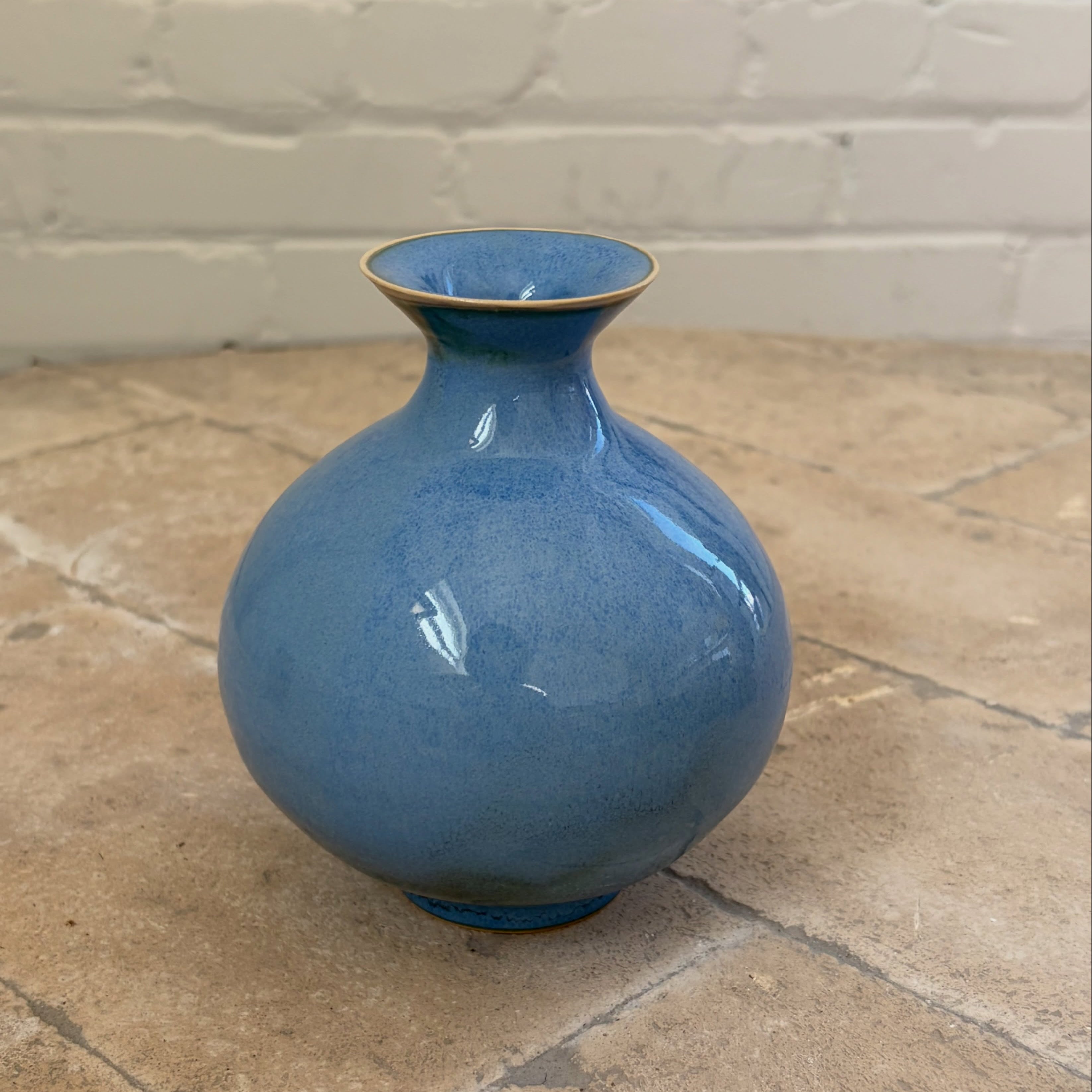 A glossy blue ceramic vase with a rounded bulbous body and a flared neck, standing on a stone-tiled floor against a white brick wall.