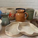 A collection of handmade ceramic tableware, including speckled beige plates, a brown speckled pitcher, a teal-green vase, and stacked ribbed bowls, arranged on a stone surface against a white brick wall.