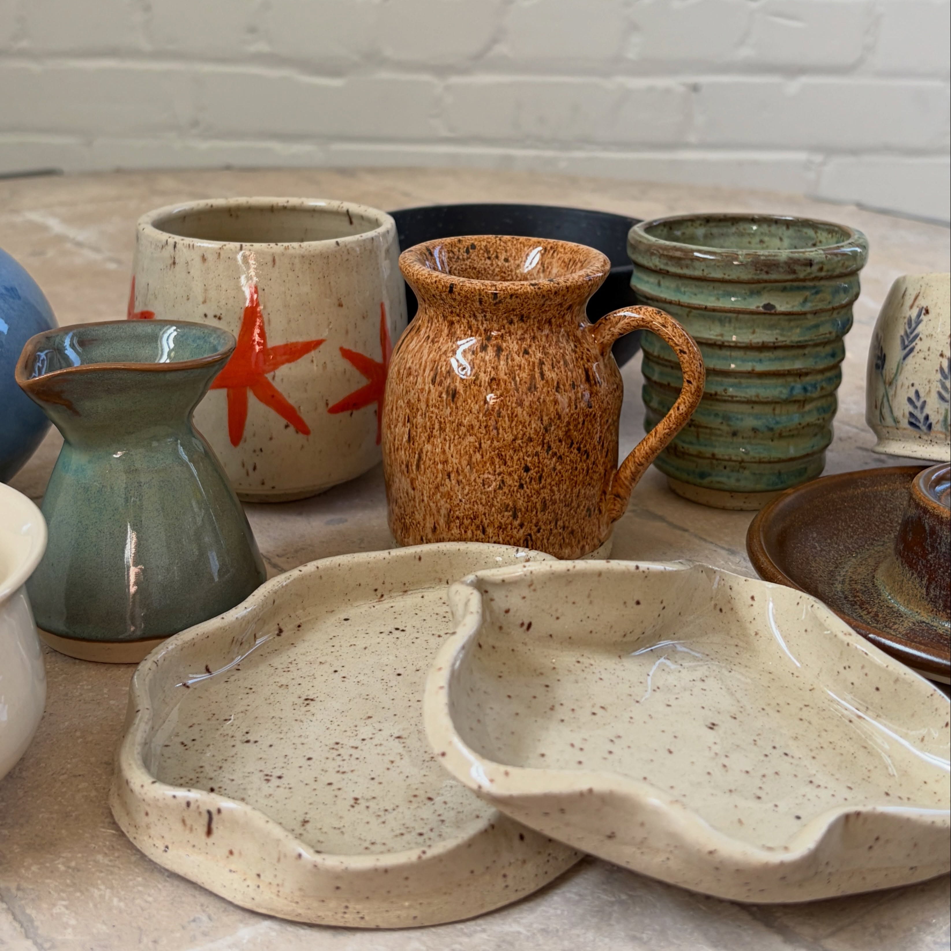 A collection of handmade ceramic tableware, including speckled beige plates, a brown speckled pitcher, a teal-green vase, and stacked ribbed bowls, arranged on a stone surface against a white brick wall.