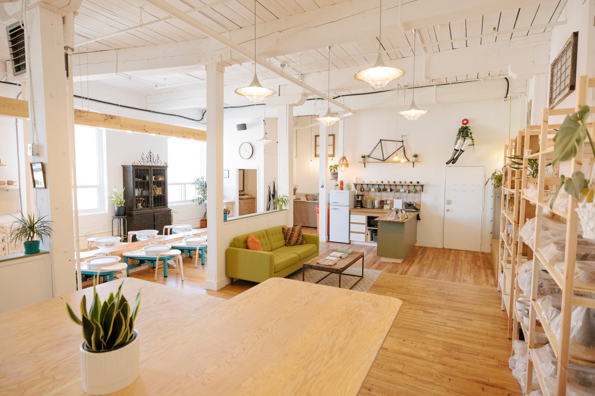 A bright, modern loft-style office or cafe interior with exposed white ceiling beams, light wood floors, pendant lights, a green sofa, a small kitchenette, shelving, and potted plants.