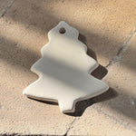 An off-white ceramic Christmas tree ornament with a hanging hole, resting on a textured stone surface in dappled sunlight.
