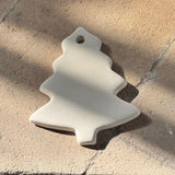 An off-white ceramic Christmas tree ornament with a hanging hole, resting on a textured stone surface in dappled sunlight.