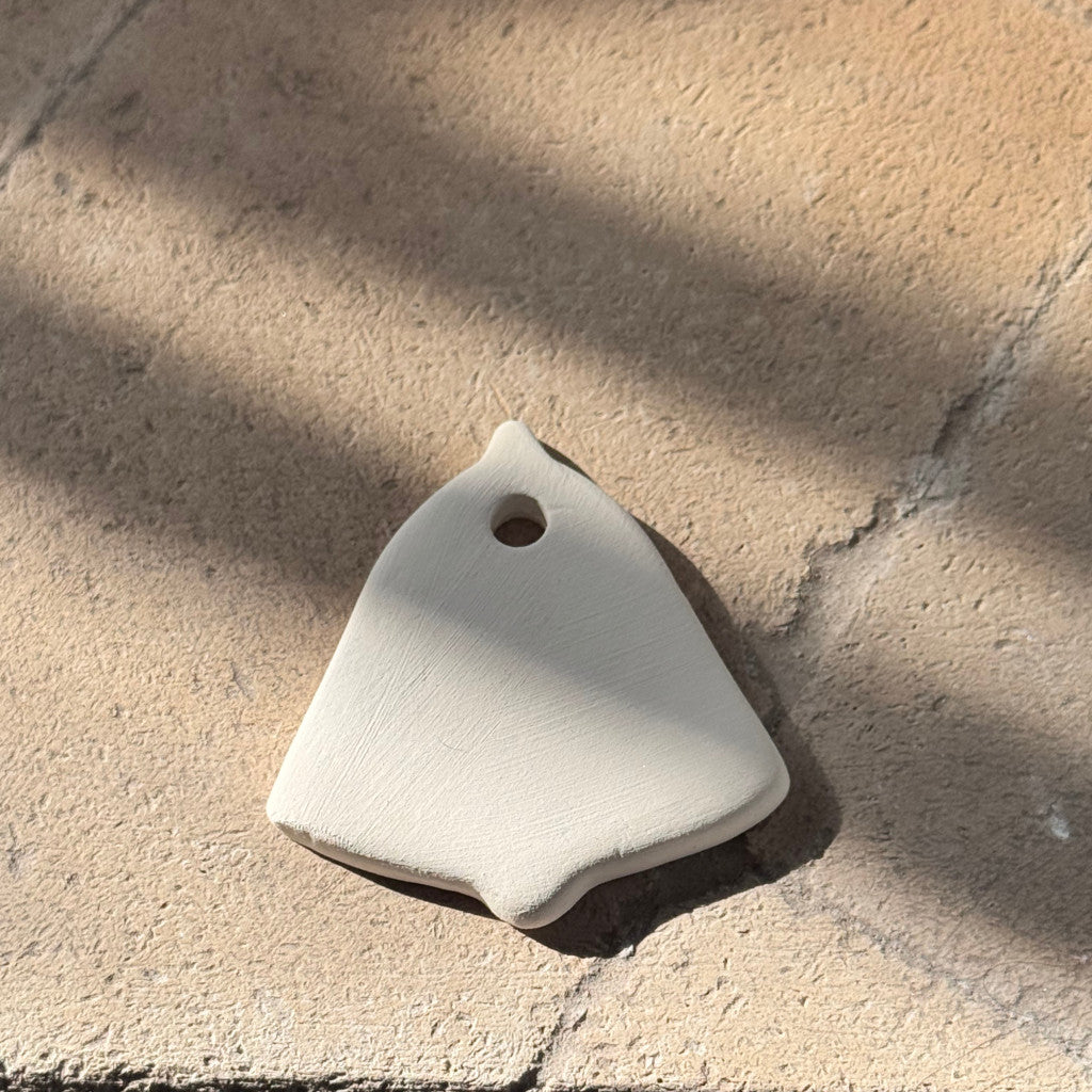 A small off-white ceramic pendant with a rounded triangular shape and a hole near the top, photographed on a textured stone surface with dappled sunlight and shadows.