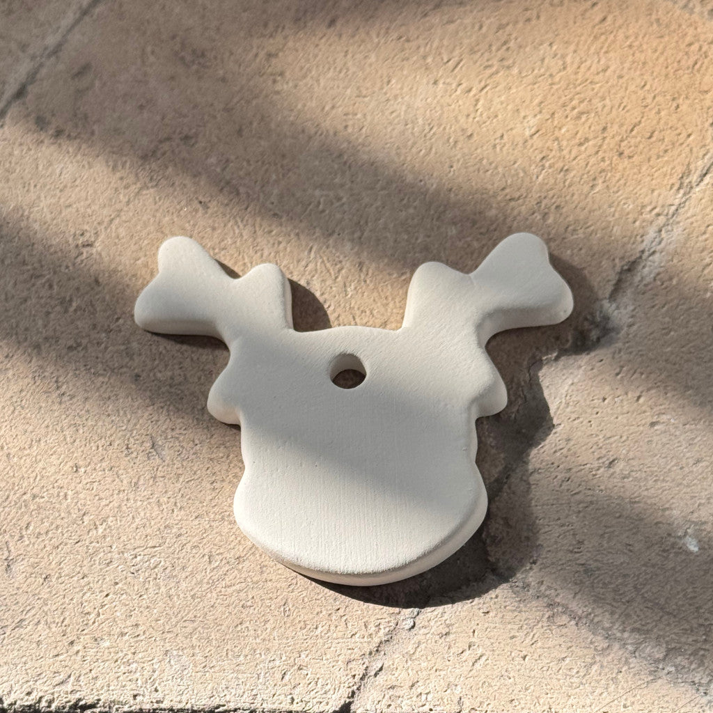 A small off-white wooden ornament shaped like a reindeer head with antlers and a hanging hole, photographed on a textured stone surface in natural sunlight.