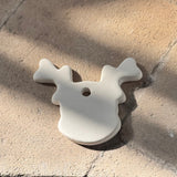 A small off-white wooden ornament shaped like a reindeer head with antlers and a hanging hole, photographed on a textured stone surface in natural sunlight.
