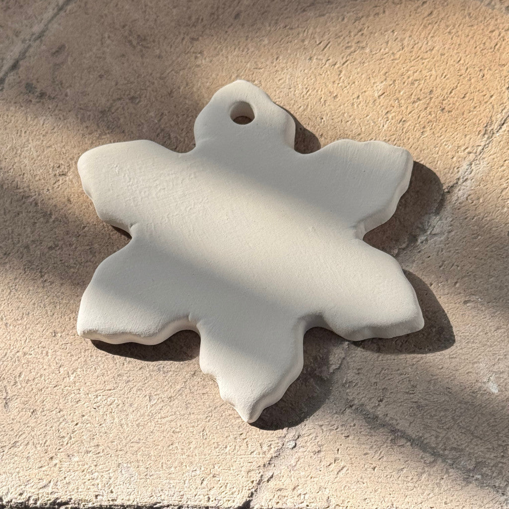 A white, snowflake-shaped ceramic charm with a small hole at the top, resting on a textured beige stone surface under direct sunlight.