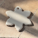 A white, snowflake-shaped ceramic charm with a small hole at the top, resting on a textured beige stone surface under direct sunlight.