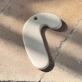 A curved, off-white ceramic wall hook with a small hole at the top, resting on a textured beige stone surface with soft shadows.